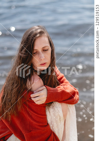 Young woman with long hair and closed eyes, in a white skirt and a terracotta sweater, sits on a tree stump against the backdrop of the sea 129519153