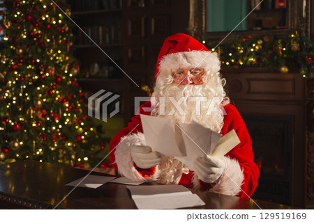 Merry Christmas. Happy Santa Claus reading letters from children in festive Christmas living room. Santa with xmas wish list letter on Christmas eve. Mail of Santa Claus. Winter Holiday Celebrations 129519169