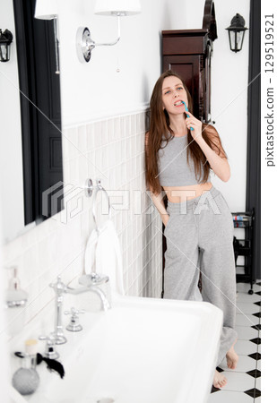 A beautiful and young woman in gray pajamas with braces stands in the bathroom leaning against the wall and brushing her teeth. A beautiful and young woman in gray pajamas with braces stands in the bathroom leaning against the wall and brushing her teeth. 129519521