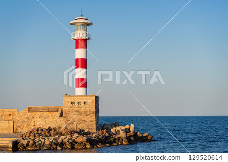 Red and white striped lighthouse on stone pier in Bulgaria Black Sea harbor during golden hour. Concept of coastal tourism, nautical navigation, iconic travel landmark 129520614