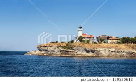 White lighthouse and traditional houses on rocky Bulgaria Black Sea coast under sunny blue sky in summer. St. Anastasia Island. Concept of travel destination, coastal landscape and maritime heritage 129520618