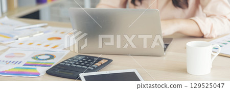 Asian Female freelance using laptop at home office desk. Woman reading financial graph chart Planning analyzing marketing data. Asian female people working office firm with business stuff, coffee cup 129525047