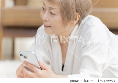 A senior woman lying on the living room floor and operating a smartphone 129526695