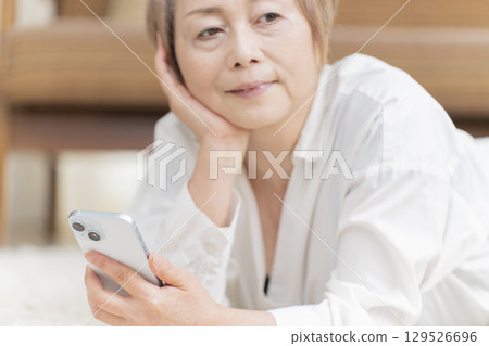 一位老年婦女躺在客廳的地板上操作智慧型手機 一位老年婦女躺在客廳的地板上操作智慧型手機 129526696