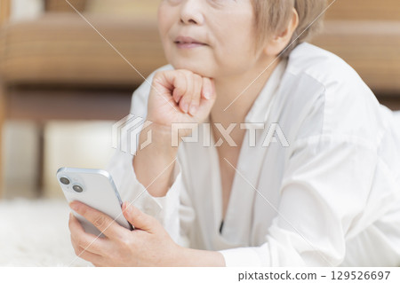 一位老年婦女躺在客廳的地板上操作智慧型手機 一位老年婦女躺在客廳的地板上操作智慧型手機 129526697