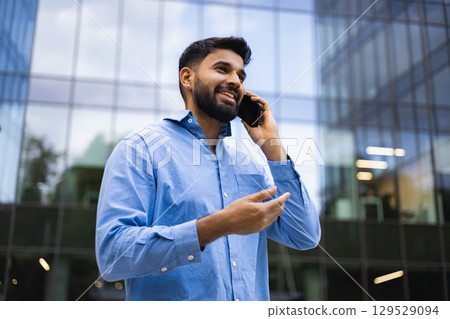 A smiling man of Indian descent is on a phone call outside of a modern building. 129529094