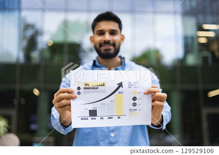 A smiling man holds a chart showing business growth outdoors with a blurred building in the background. A smiling man holds a chart showing business growth outdoors with a blurred building in the background. 129529106