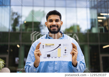 A man in a blue shirt smiles, holding a paper with business charts in front of a modern building. A man in a blue shirt smiles, holding a paper with business charts in front of a modern building. 129529107