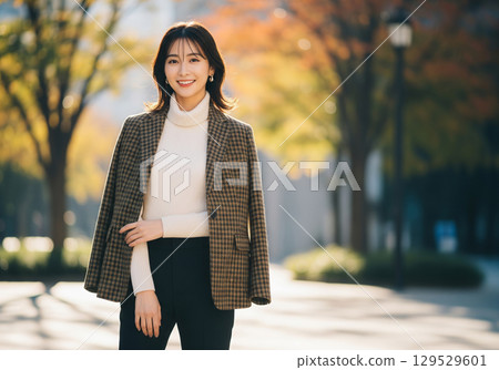 A woman smiling on an elegant autumn tree-lined street wearing a checked jacket draped over her shoulders (autumn outfit) 129529601
