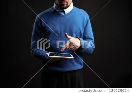 Man in blue sweater using tablet while standing against a dark background in a modern setting Man in blue sweater using tablet while standing against a dark background in a modern setting 129529624