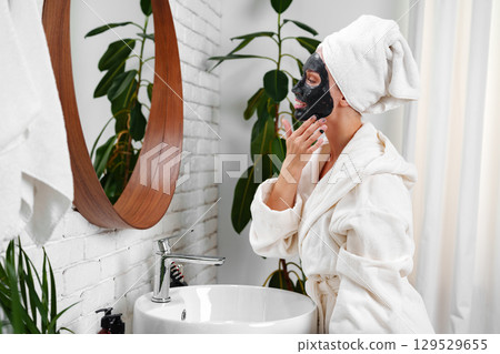 Woman applying face mask in bathroom while wearing a bathrobe and towel, surrounded by indoor plants Woman applying face mask in bathroom while wearing a bathrobe and towel, surrounded by indoor plants 129529655