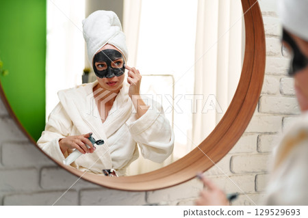 Woman applies facial mask while wearing a robe in a well-lit bathroom with a large round mirror 129529693