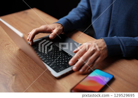 Hands typing on a laptop keyboard with a smartphone resting on a wooden desk during the evening 129529695