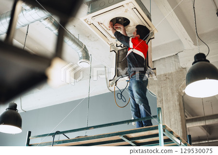 Technician performs installation work on ceiling-mounted air conditioning unit in modern office Technician performs installation work on ceiling-mounted air conditioning unit in modern office 129530075