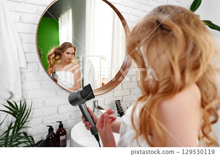 Woman applying skincare while looking in mirror after shower in bright bathroom 129530119