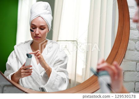 Woman applying skincare product while wearing a robe and towel in a bright bathroom setting 129530178