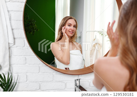 Woman applying skincare product in a bright bathroom with green accents and natural light 129530181