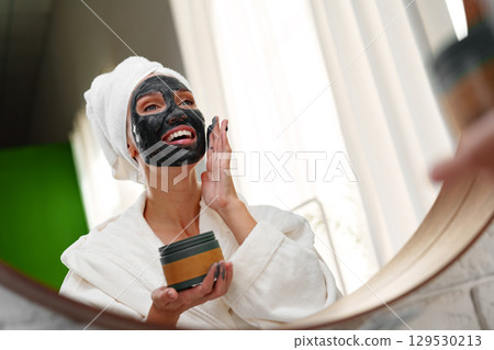 Woman applying facial mask while smiling in a bright indoor space during a self-care routine 129530213