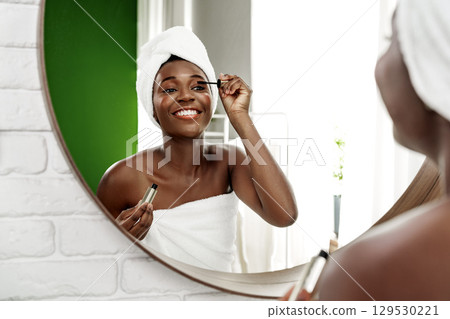 Woman applying mascara while smiling in a bright bathroom with green accents and a large mirror Woman applying mascara while smiling in a bright bathroom with green accents and a large mirror 129530221