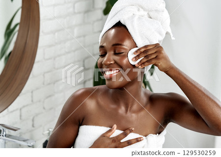 Smiling woman enjoying skincare routine in a bright bathroom with a towel on her head during the daytime 129530259