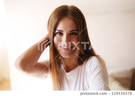Young woman with long brown hair smiling and posing casually in a bright, cozy indoor setting Young woman with long brown hair smiling and posing casually in a bright, cozy indoor setting 129530378
