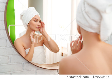 Woman applying skincare while looking in mirror after shower in bright bathroom Woman applying skincare while looking in mirror after shower in bright bathroom 129530383