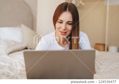 Woman using laptop while sitting on bed in cozy bedroom during daytime Woman using laptop while sitting on bed in cozy bedroom during daytime 129530477