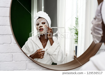 Self-care routine in a bright bathroom with a woman applying a facial mask and wearing a robe 129530517