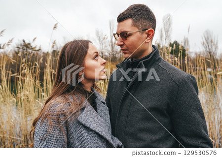 Couple gazes at each other amid tall grass by the lake during a cloudy day in autumn 129530576