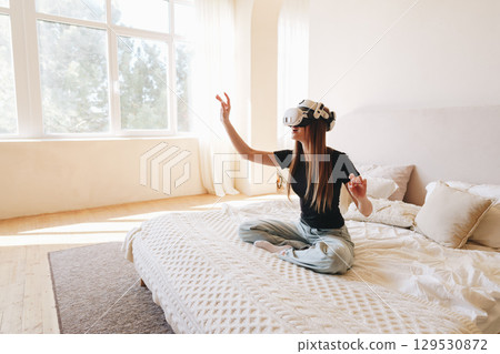 Young woman enjoys virtual reality experience while sitting on a bed in a bright room during daytime Young woman enjoys virtual reality experience while sitting on a bed in a bright room during daytime 129530872