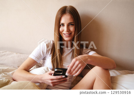 Young woman sitting on a bed, smiling and using a smartphone in a cozy room during daytime for a casual moment of relaxation 129530916