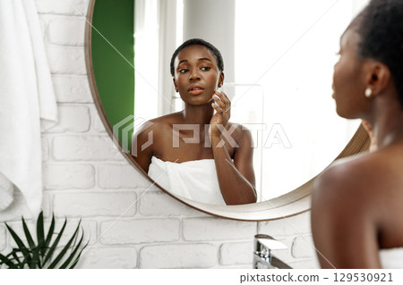 Woman applying skincare in a modern bathroom mirror during morning routine for glowing complexion 129530921
