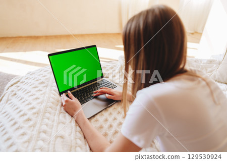 Woman sits on a cozy bed working on a laptop with a green screen during daylight in a bright room 129530924