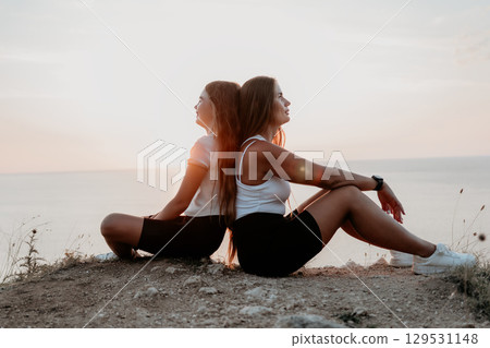 Two Women Sitting Back to Back on a Cliffside at Sunset 129531148
