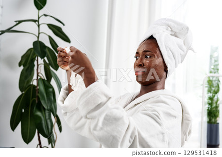 Woman in a towel applying skincare treatment at home in a bright, modern bathroom with indoor plants 129531418