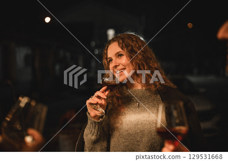 Woman smiling while holding a glass of wine at a nighttime gathering with friends outdoors 129531668