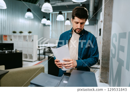 Man using a printer in a modern office space during the day while reviewing printed documents Man using a printer in a modern office space during the day while reviewing printed documents 129531843