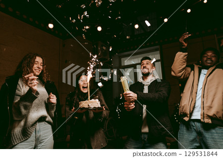 Friends celebrating a birthday with cake, sparkles, and joyful expressions in an indoor party setting 129531844