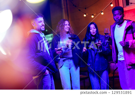 Friends enjoy an evening gathering with drinks under colorful lights in an outdoor setting Friends enjoy an evening gathering with drinks under colorful lights in an outdoor setting 129531891