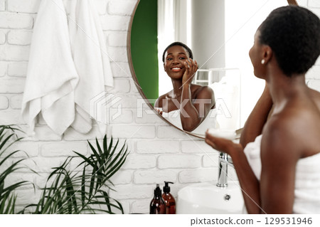 Woman applying skincare in a modern bathroom with green accent and natural light Woman applying skincare in a modern bathroom with green accent and natural light 129531946