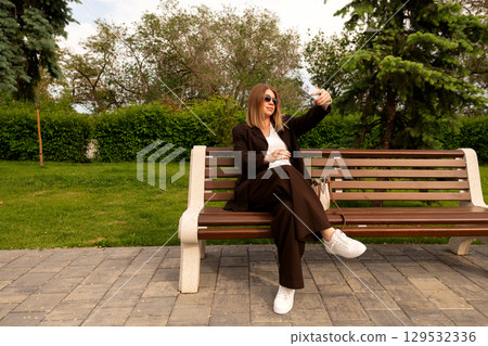 Young stylish woman takes selfie while seated on bench outdoors. Young stylish woman takes selfie while seated on bench outdoors. 129532336