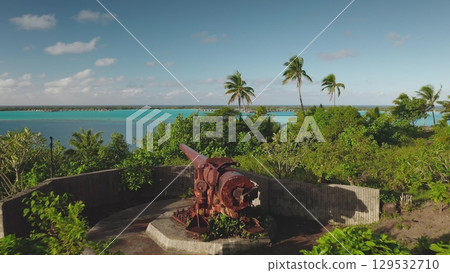 Lush vegetation covers a tropical island, where a rusty cannon overlooks turquoise waters, small boats dotting the tranquil lagoon, with palm trees swaying gently in the breeze Lush vegetation covers a tropical island, where a rusty cannon overlooks turquoise waters, small boats dotting the tranquil lagoon, with palm trees swaying gently in the breeze 129532710