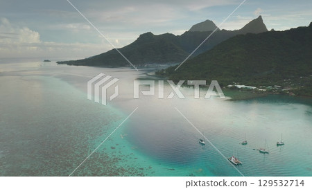 Aerial view of sailboats moored in turquoise lagoon near a lush tropical island with volcanic mountains, creating a picturesque scene of exotic paradise. Wild travel destination, remote summer nature Aerial view of sailboats moored in turquoise lagoon near a lush tropical island with volcanic mountains, creating a picturesque scene of exotic paradise. Wild travel destination, remote summer nature 129532714