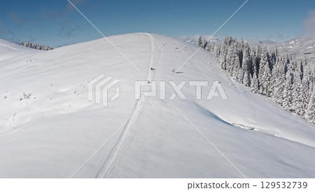 Two snowmobiles driving up a snowy mountain slope, leaving tracks in fresh snow, on a sunny day with clear blue sky and snowy forest on the side. Bukovel ski resort. Winter nature travel background Two snowmobiles driving up a snowy mountain slope, leaving tracks in fresh snow, on a sunny day with clear blue sky and snowy forest on the side. Bukovel ski resort. Winter nature travel background 129532739