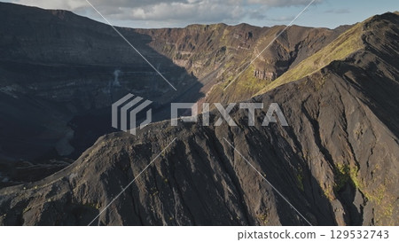 Drone captures rugged beauty of a volcanic crater, smoke and steam rising from its depths as clouds drift across sky, casting shadows on dramatic landscape. Amazing nature travel background 129532743