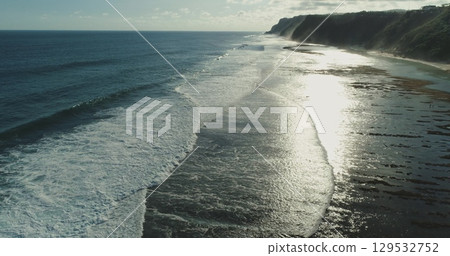 Sunlight shimmering on the shallow waters along Bali's coast, with gentle waves rolling toward the shore as the sun sets, creating a serene and picturesque tropical landscape 129532752