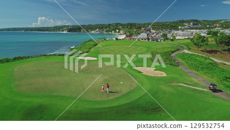 Two golfers are playing on a lush green golf course overlooking the ocean in Bali, Indonesia, enjoying the stunning views and tropical weather 129532754