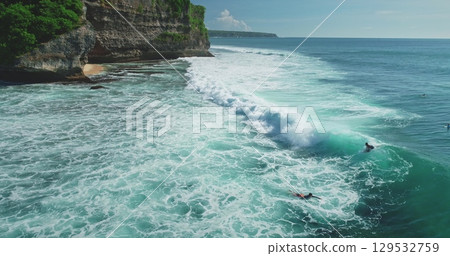 Turquoise ocean waves rolling towards the shore at Pantai Dreamland Beach, Bali, with surfers riding the waves and cliffs in the background on a sunny day 129532759