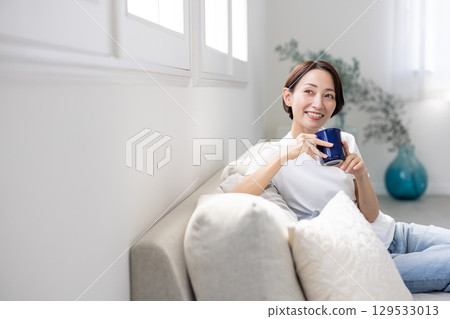A woman sitting on a sofa looking out the window with a blue mug in her hand 129533013