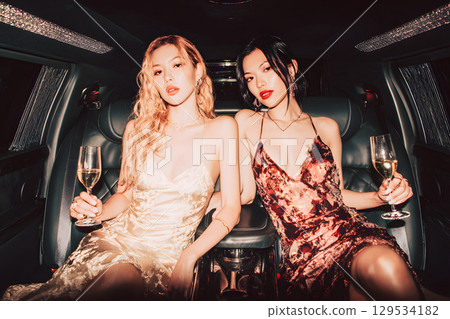 兩位年輕、名媛般的女性在豪華轎車裡舉杯慶祝,度過了一個迷人的夜晚 兩位年輕、名媛般的女性在豪華轎車裡舉杯慶祝,度過了一個迷人的夜晚 129534182